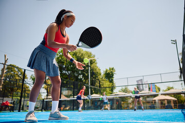 Black woman serving ball while playing paddle tennis doubles on court.