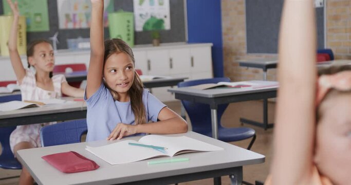 Happy diverse schoolgirls raising hands at desks in elementary school class, slow motion