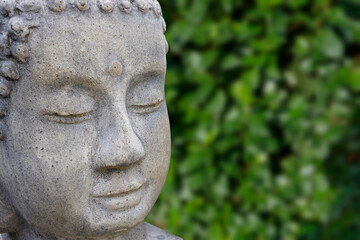Close-up of a Buddha head made of gray stone. In the background, green blurred plants.