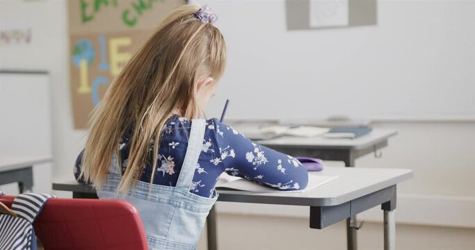 Rear view of caucasian schoolgirl writing at desk in elementary school class, slow motion