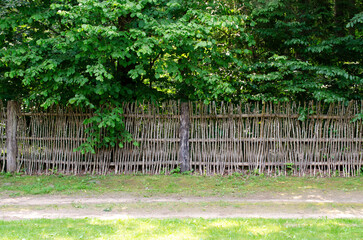 Wattle is a lightweight building material made by interlacing branches. Wattle fence against the background of a green forest, reserve. Wicker fence made of branches, rustic fence.