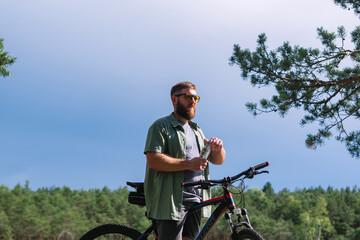 Cyclist have rest near the bicycle, in the forest and relax for energy in adventure workout, drinking bottle of water for hydration and  enjoying in the forest trees for wellness. Bearded man on bike 