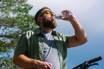 Cyclist have rest near the bicycle, in the forest and relax for energy in adventure workout, drinking bottle of water for hydration and  enjoying in the forest trees for wellness. Bearded man on bike 