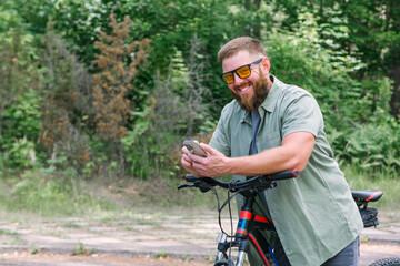 Happy bearded man in jeans clothes stand near bicycle bike on sidewalk in green park outdoors use mobile cell phone 13 Pro chat online. People active urban healthy lifestyle cycling concept