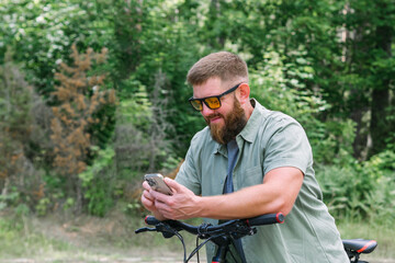 Happy bearded man in jeans clothes stand near bicycle bike on sidewalk in green park outdoors use mobile cell phone 13 Pro chat online. People active urban healthy lifestyle cycling concept