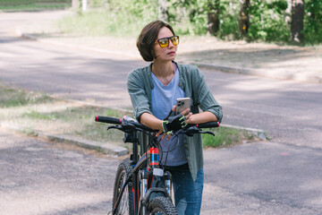 Happy woman in jeans clothes stand near bicycle bike on sidewalk in green park outdoors use mobile cell phone 13 Pro chat online. People active urban healthy lifestyle cycling concept