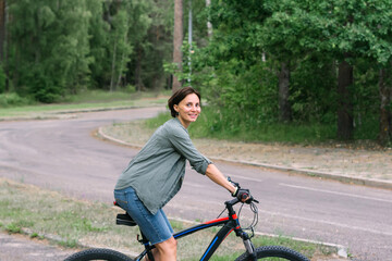 Satisfied smiling happy woman in casual green jacket jeans riding bicycle bike on sidewalk in park outdoors, look camera. People active urban healthy lifestyle cycling concept.