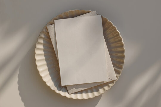Summer greeting card mockup. Blank wedding invitation, template with craft envelope on beige ceramic scallop dinner plate in sunlight. Beige table background. Minimal composition. Flatlay, top view.
