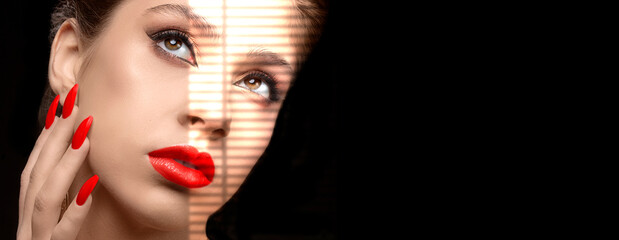 A model with red lipstick and nail polish is illuminated by the bright light of the blinds on a...