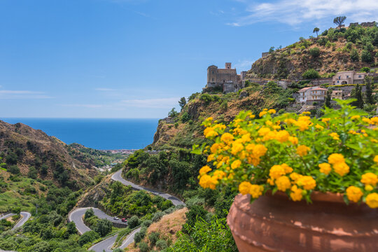 Picturesque view a Church of San Nicolo in Savoca village in Sicily, Italy. Houses on the hill  and winding road. Defocused vase with yellow flowers in the foreground. 
