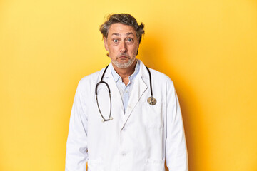 Middle-aged doctor standing against a yellow backdrop shrugs shoulders and open eyes confused.