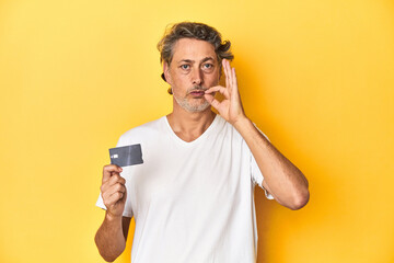 Man holding a credit card, yellow studio backdrop with fingers on lips keeping a secret.