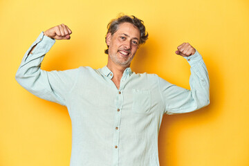 Fototapeta premium Middle-aged man posing on a yellow backdrop showing strength gesture with arms, symbol of feminine power