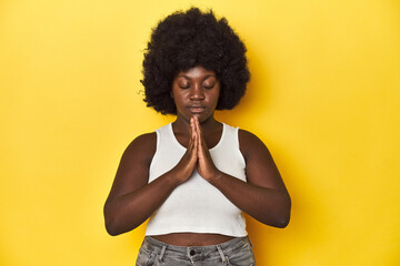 Fototapeta na wymiar African-American woman with afro, studio yellow background holding hands in pray near mouth, feels confident.