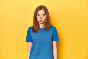 Redhead in blue t-shirt on yellow backdrop blows cheeks, has tired expression. Facial expression...