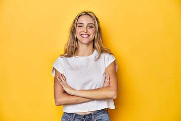 Young blonde Caucasian woman in a white t-shirt on a yellow studio background, who feels confident, crossing arms with determination. - obrazy, fototapety, plakaty