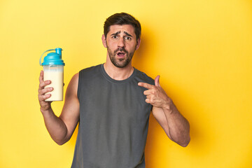 Fit man with protein shake, yellow studio background pointing to the side