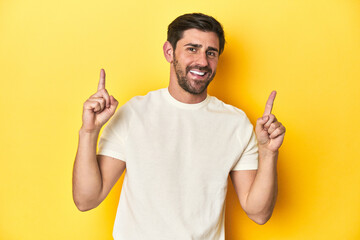 Caucasian man in white t-shirt on yellow studio background indicates with both fore fingers up...