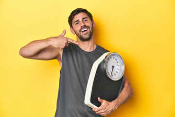Sporty Caucasian man hugging a scale on a yellow studio background.