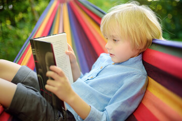 Cute little blond caucasian boy reading book and having fun with multicolored hammock in backyard...
