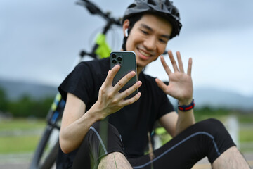 Fototapeta premium Smiling asian man cyclist taking a break resting from cycling bike in the morning and using mobile phone