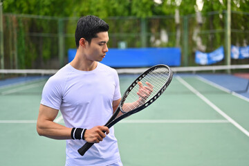 Portrait of asian male tennis player with racket standing at court on sunny day. Sport, training and active life concept