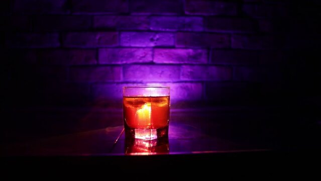 Whiskey drink concept. Glass of whiskey and ice with color light and fog on dark bar background. Selective focus