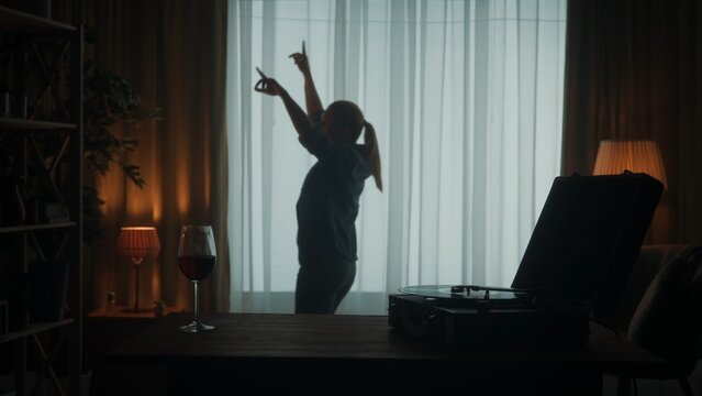 Vintage record player with a vinyl record and a glass of red wine on the table close up. Carefree woman dances cheerfully alone near the window. Dancing woman silhouette in the living room.