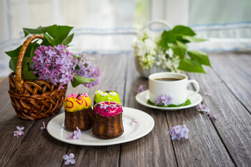 Cupcakes with colorful decor on a white plate, lilacs in a basket and a cup of hot coffee on a wooden background.
