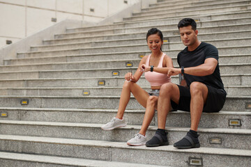 Fototapeta na wymiar Smiling couple sitting on steps and checking fitness trackers after morning jog