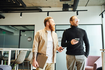 businessman in black turtleneck and eyeglasses standing with hand in pocket and looking away near bearded colleague with laptop and smartphone in modern office, productivity and cooperation concept