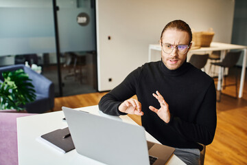 serious entrepreneur in black turtleneck and eyeglasses gesturing and talking while having video call on computer in contemporary office space, business lifestyle concept