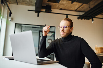 thoughtful entrepreneur in black turtleneck and eyeglasses holding pen and looking at laptop while...