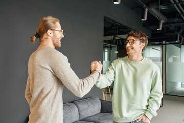 cheerful and stylish entrepreneurs in eyeglasses and casual clothes shaking hands and confirming agreement in lounge of modern coworking office, successful collaboration concept