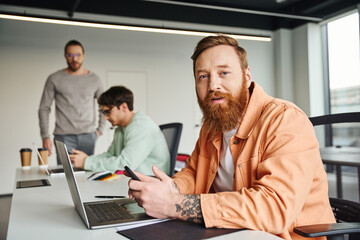 bearded and tattooed businessman in stylish casual clothes holding smartphone and looking at camera near laptop and colleagues working on blurred background in contemporary coworking space