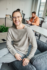 happy entrepreneur in stylish eyeglasses looking at camera in modern coworking environment near colleagues working on blurred background, successful entrepreneurship concept