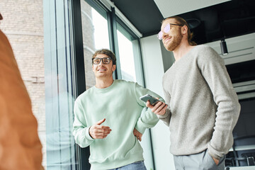Fototapeta premium smiling entrepreneur in stylish eyeglasses holding mobile phone while standing with hand in pocket near business team and large windows in coworking office, concept of successful collaboration
