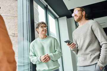 businessman in eyeglasses and stylish casual clothes standing with hand in pocket and smartphone near happy colleague and large window in modern office, concept of successful collaboration