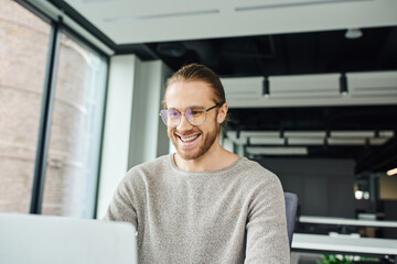 cheerful and successful entrepreneur in casual clothes and stylish eyeglasses looking at laptop while working on startup project in contemporary office, business inspiration concept