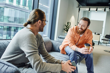cheerful, bearded and tattooed businessman with coffee to go discussing startup project with colleague sitting with mobile phone on couch in office lounge of modern coworking environment