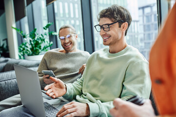 happy entrepreneur in eyeglasses pointing at laptop while working on startup project near business...
