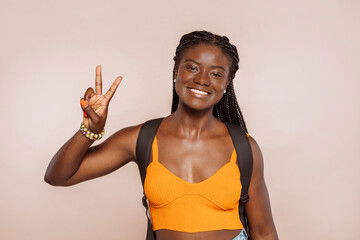 Young african female student, wearing casual orange top and backpack, gesturing peace victory sign...