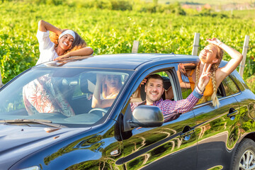 Happy friends waving hands outside open window car during road trip with vineyards in background - 621851409