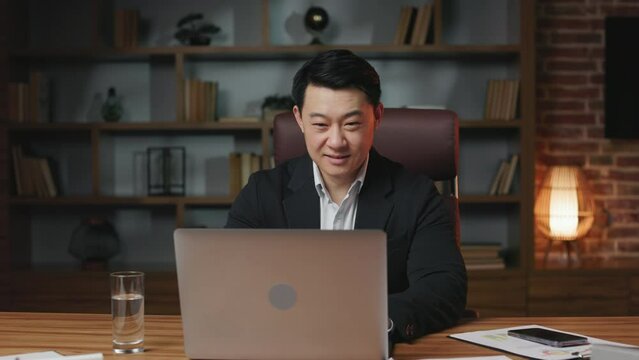 Positive asian gentleman with dark hair writing few last sentences in financial report and closing laptop. Cheerful employee dressed in business clothes ending workday and leaving office.