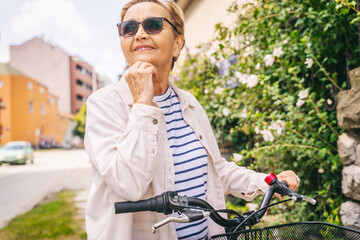 Attractive happy senior woman in sunglasses standing with bicycle in sunny summer city