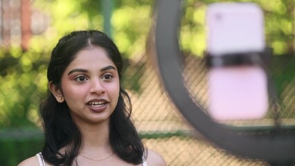 Cheerful young indian woman influencer recording video blog mobile vlog podcast live stream share experience with audience on smartphone using tripod at green park