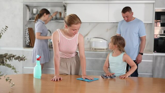 Joint family house cleaning, household tasks, household help. Eldest girl wipe dry kitchen table, father with little daughter vacuums floor in background, mother mop dry dishes