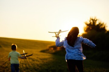 Running boy and girl holding two yellow and blue airplanes toy in the field during summer sunset. Kids dreams of flying and aviation.