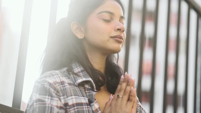 Portrait of hopeful young indian woman clasping hands in prayer asking for blessing and help while the rays of rising sun fall on her face near church outdoors Religion and faith concept	