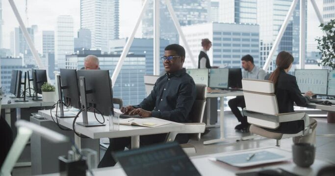 Focused African American Male Data Analyst Typing On Desktop Computer In Diverse Corporate Office With Megapolis Window View. Professional Balck Man Working In Consulting Company With Colleagues.